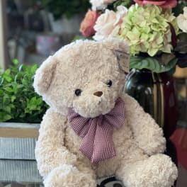 Plush teddy bear with a plaid bow sitting beside a floral arrangement