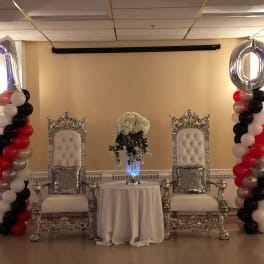 Balloon columns flank two ornate chairs with a floral centerpiece between them.