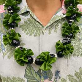 Glossy black kukui nut lei with green ribbon clusters worn over a tropical shirt.