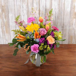 Colorful mixed bouquet with roses, gerbera daisies, lilies, and tulips in a round glass vase with striped ribbon.
