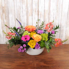 Low oval arrangement of bright orange, yellow, pink, and purple flowers in a white ceramic vase.