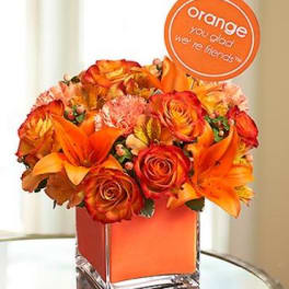 Orange roses and lilies arranged in a square glass vase with a round sign