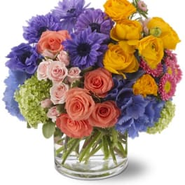 Mixed bouquet of purple, yellow, pink, and blue flowers in a clear glass vase