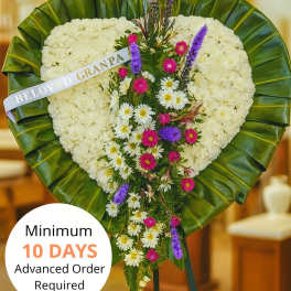 Heart-shaped white sympathy standing spray with cascading purple and pink flowers on a green-leaf border