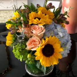 Bouquet of sunflowers, pale roses, blue hydrangea, and green blooms in a glass vase