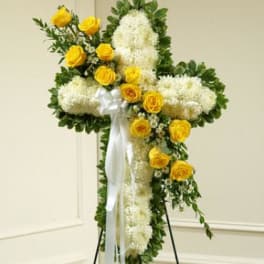 Cross-shaped floral tribute with white blooms and yellow roses on a stand