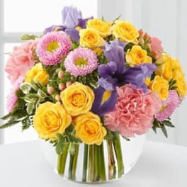 Mixed bouquet of yellow roses, purple irises, pink carnations, and pink mums in a glass vase