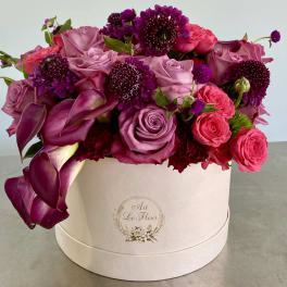 Pink and purple roses and calla lilies arranged densely in a round ivory hatbox container.