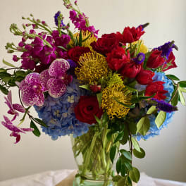 Tall mixed arrangement of red roses, yellow pincushion protea, blue hydrangeas, and spotted orchids in a clear glass vase