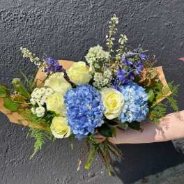 Hand-tied bouquet of blue hydrangeas, cream roses, and white daisies wrapped in kraft paper.