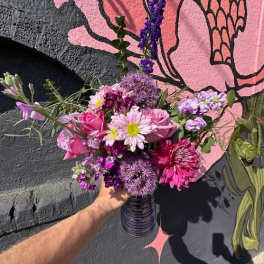 Hand holding a bright pink and purple mixed flower bouquet in a ribbed purple glass vase.