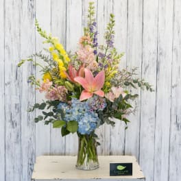 Mixed bouquet with pink lilies, blue hydrangea, and tall yellow and purple blooms in a glass vase