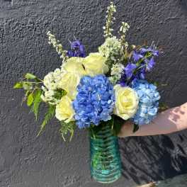 Arrangement of yellow roses, blue hydrangeas, and white flowers in a teal glass vase