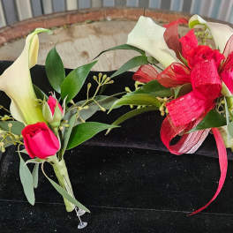 Two calla lily corsages with red ribbon and pink rosebuds on a black box.