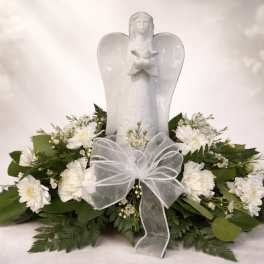 White angel figurine surrounded by white flowers and greenery with a sheer white bow.