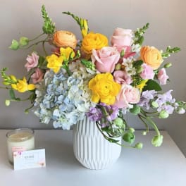 Mixed pastel bouquet in a white vase with a candle and card beside it