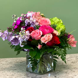 Mixed arrangement of pink and red roses with purple flowers in a clear glass cube vase