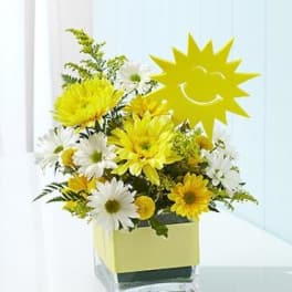 Yellow and white daisy bouquet in a square glass vase with a sun pick