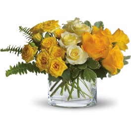Low arrangement of yellow and white roses in a clear glass cube vase with fern accents