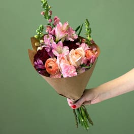 Hand-tied bouquet of pink and peach roses, ranunculus, and mixed blooms wrapped in brown paper.