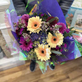 Hand-tied bouquet of yellow gerbera daisies and purple flowers wrapped in iridescent cellophane.