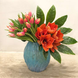 Asymmetrical vase arrangement with pink tulips, orange blooms, and a fan of glossy green leaves in a blue vase.