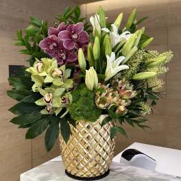Tall arrangement of orchids, lilies and protea in a textured gold vase on a marble counter