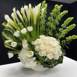 Tall white arrangement of calla lilies, roses, tulips and hydrangea in a low white bowl with green accents