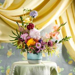 Colorful mixed flower arrangement in a blue ceramic vase on a draped table with yellow fabric backdrop.