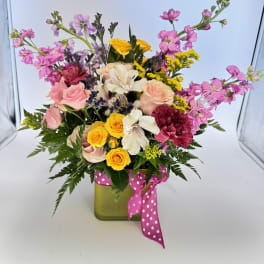 Colorful mixed flower arrangement in a green cube vase with a pink polka dot ribbon.