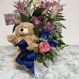 Pink lilies and roses with blue roses in a white container alongside a teddy bear and blue bow.
