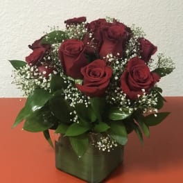 Compact arrangement of red roses with white filler flowers in a green cube vase on an orange surface