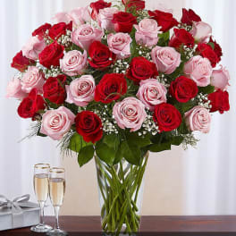 Tall clear vase of red and pink roses with baby's breath, next to two champagne glasses and a silver gift box.