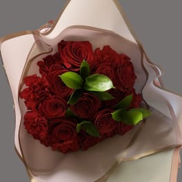 Hand-tied bouquet of red roses and red carnations with bright green leaves wrapped in white paper.