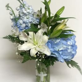 Tall glass vase with blue hydrangeas, white lilies, and other white flowers with a pearl cross on the vase