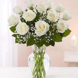 Arrangement of white roses with small white filler flowers in a clear glass vase.