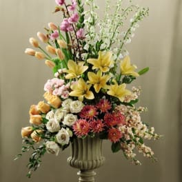 Tall urn arrangement with yellow lilies, peach tulips, roses, and mixed pastel flowers on a pedestal