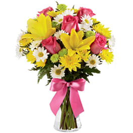 Mixed bouquet of yellow lilies, pink roses, white daisies, and mums in a clear vase with a pink bow