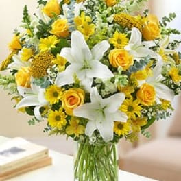 Arrangement of white lilies, yellow roses, and yellow daisies in a clear glass vase