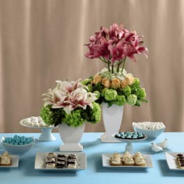 Dessert table with two pink and green floral arrangements in white vases surrounded by small pastries and candies.