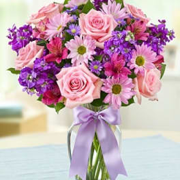 Pink roses and purple daisies in a glass vase with a lavender ribbon.