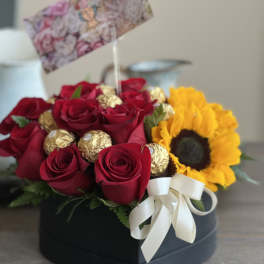 Red roses and a sunflower in a black box with gold chocolates and a white ribbon