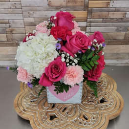 Compact arrangement of hot pink roses, white hydrangea, and pink carnations in a heart-print cube container.