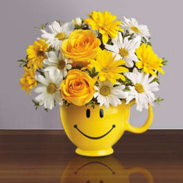 Yellow roses and white and yellow daisies arranged in a yellow smiley-face mug on a table