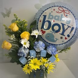 Baby boy-themed bouquet with yellow roses, blue carnations, daisies, and a baby boy balloon in a green cube vase