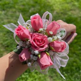 Pink rose wrist corsage with white filler flowers, ribbon loops, and bead accents on a person's wrist