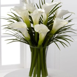 Tall arrangement of white calla lilies with long blades of foliage in a clear glass vase