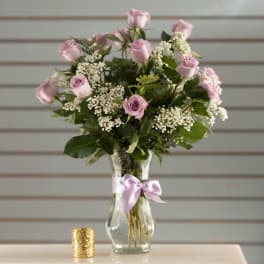 Tall arrangement of lavender roses and white filler flowers in a clear vase with a bow