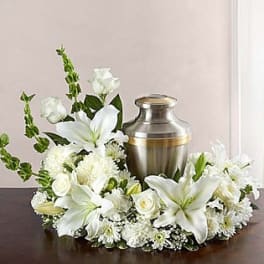 White lilies, roses, and mums arranged in a ring around a silver urn on a dark table.