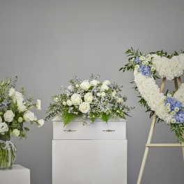 Sympathy set with white and blue arrangements and a heart-shaped wreath beside a white casket.